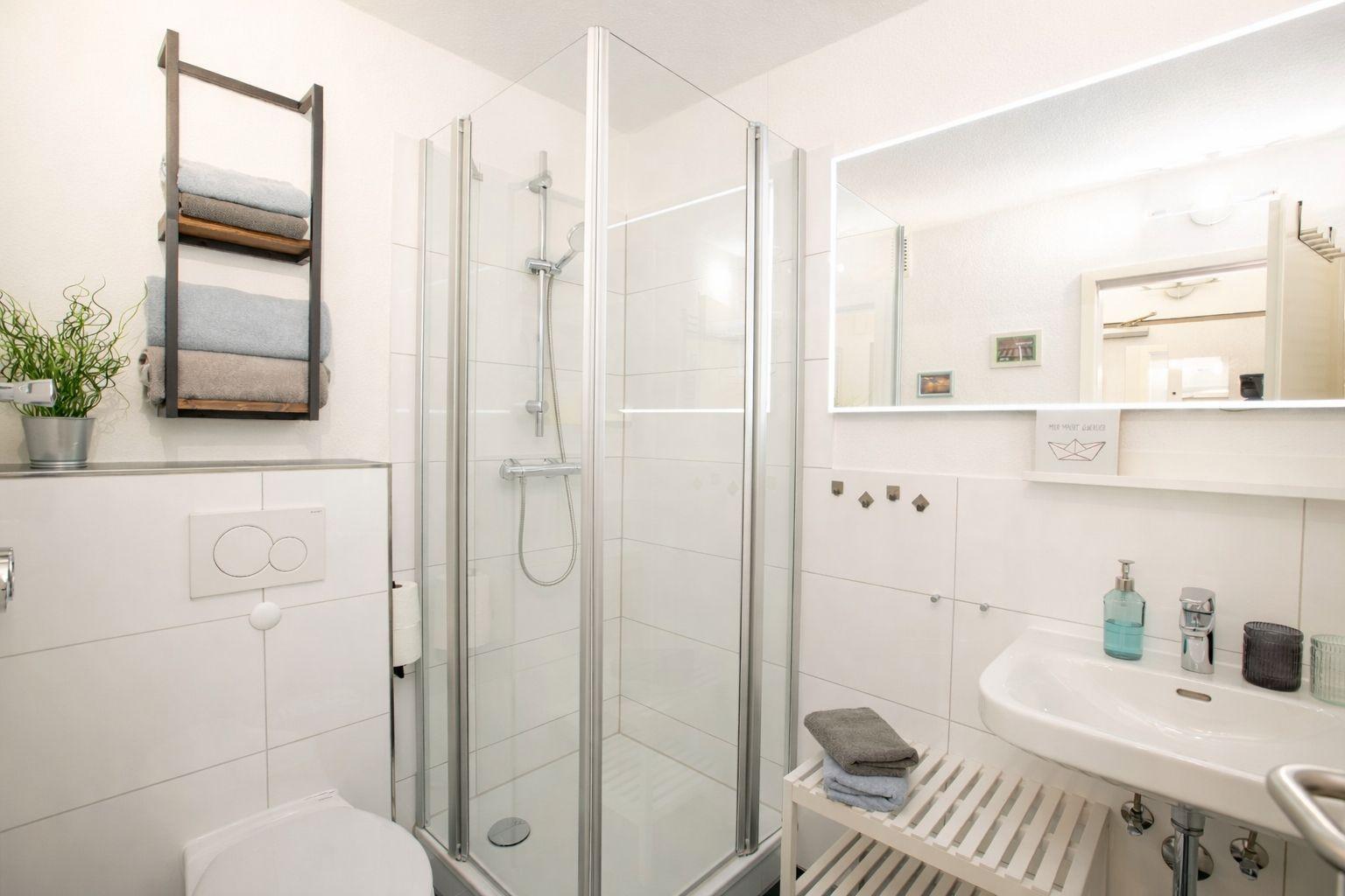 Badezimmer mit Dusche, Waschbecken und Toilette. Handtücher und Pflanze sind sichtbar.