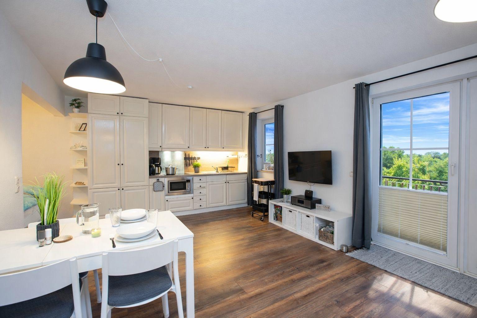 Küche mit Esstisch, Wohnbereich mit TV und Balkon mit Blick auf Grün.