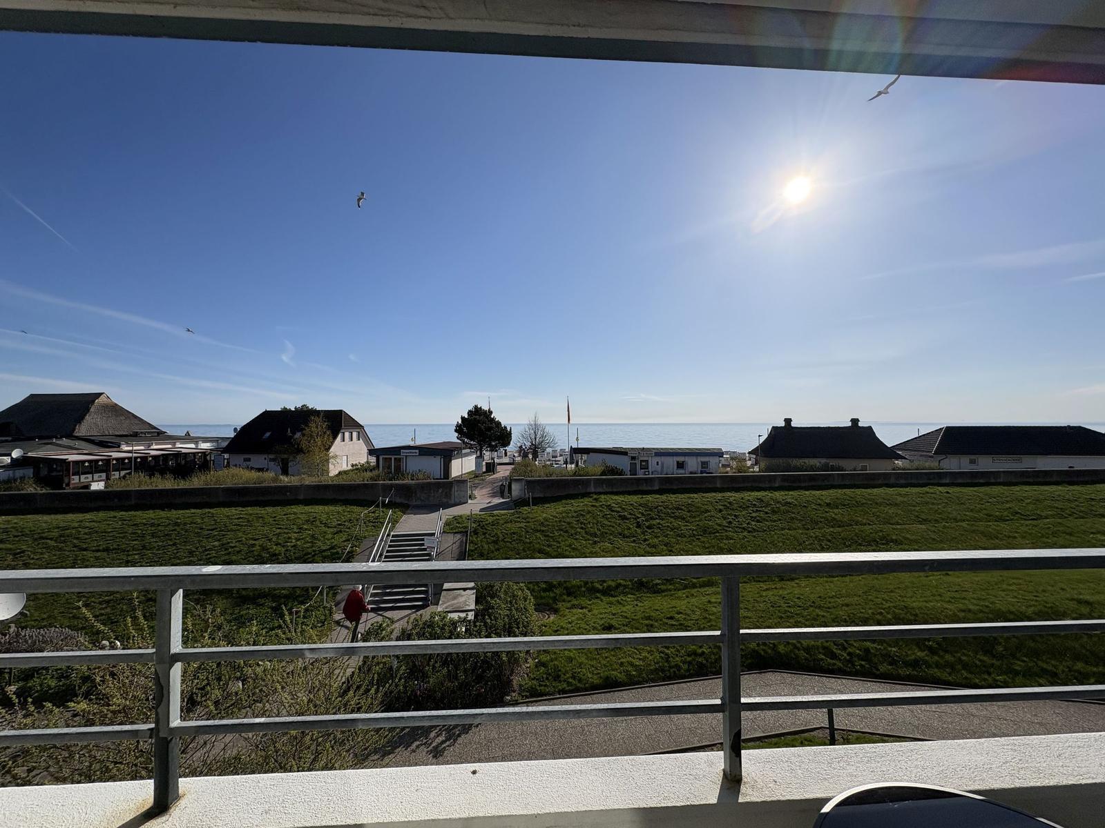 Blick vom Balkon auf Grünfläche, Häuser und Meer unter blauem Himmel mit Sonne und Möwen.