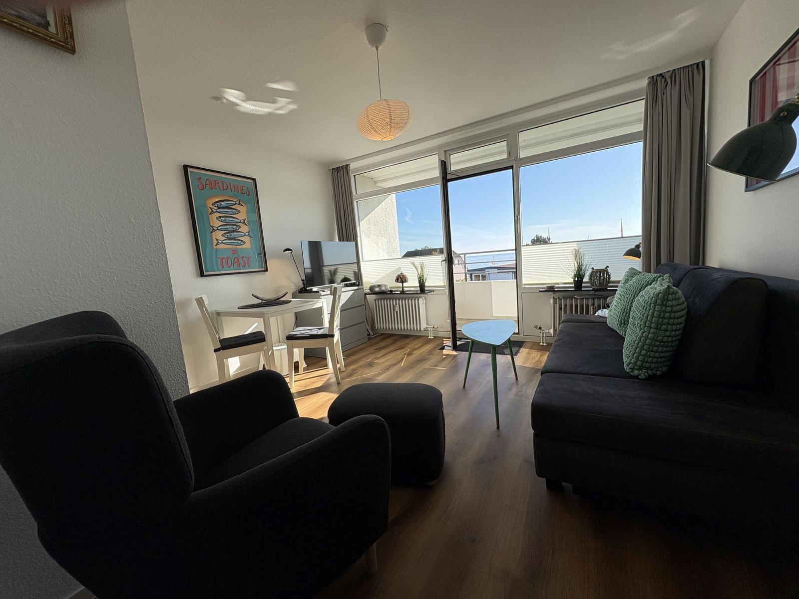 Wohnzimmer mit Sofa, Sessel und Balkonansicht. Holzboden und große Fenster.