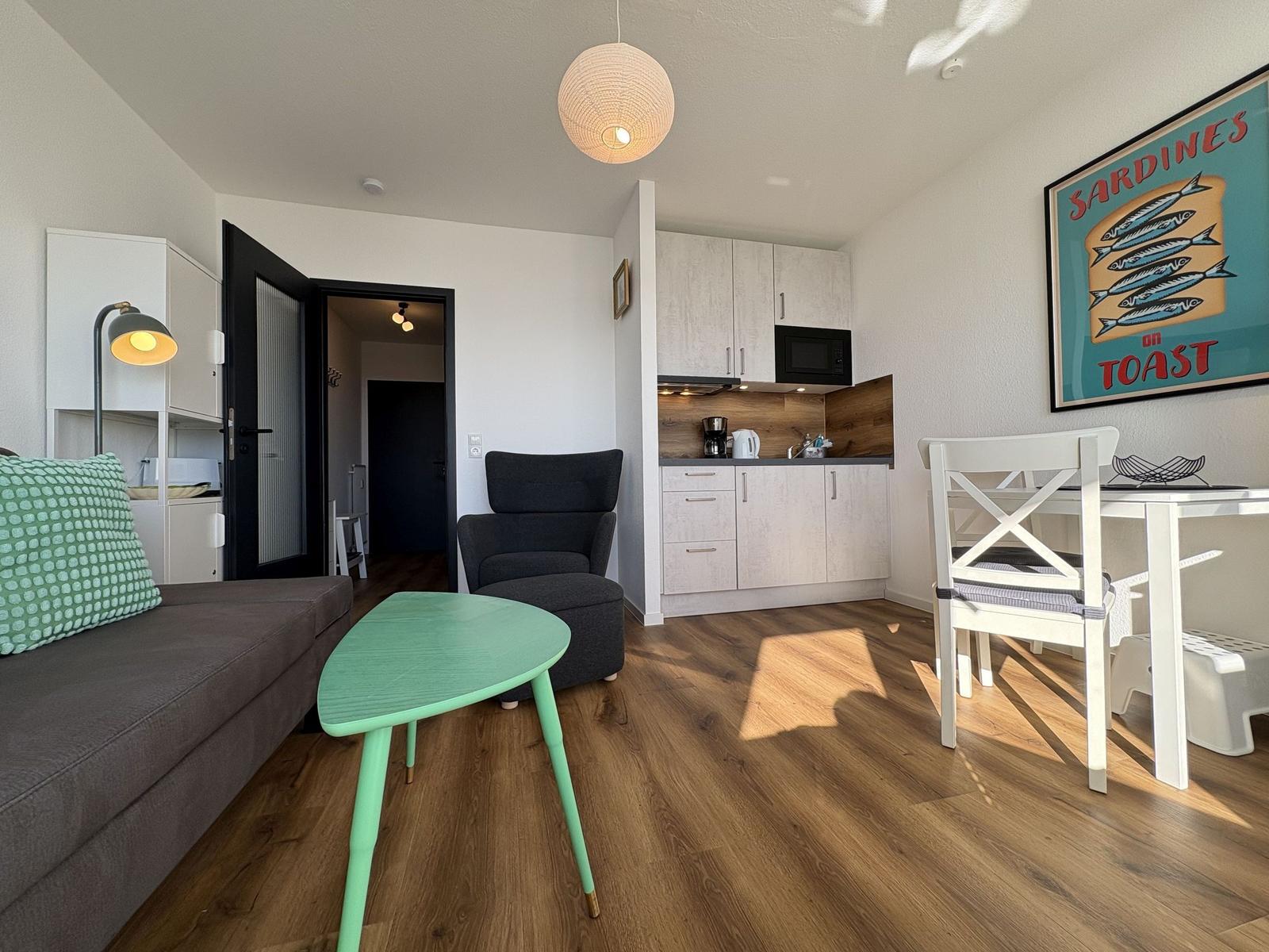 Wohnbereich mit Sofa, Esstisch und Küchenzeile. Holzboden und Wandbild sichtbar.