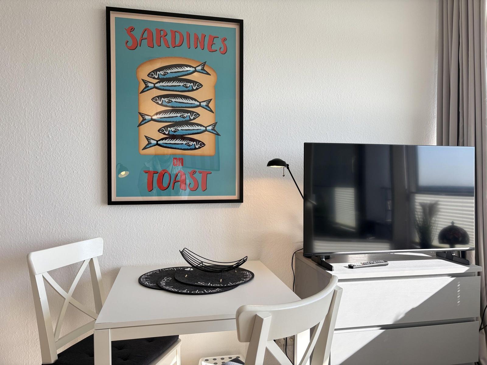 Tisch mit zwei Stühlen, Fernseher und Wandbild mit Sardinenmotiv.