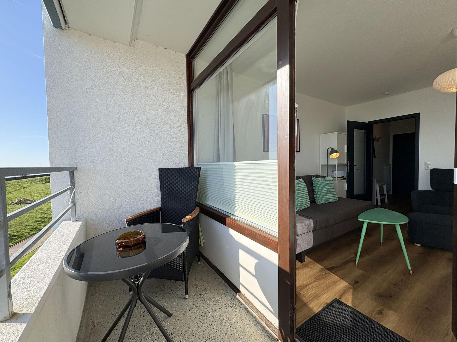 Balcony with table and chair overlooks green landscape. Living room with sofa, green cushions, and wooden floor.
