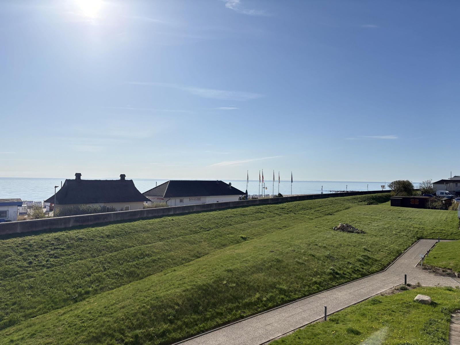 Grüner Hang mit Weg, Blick auf Strandhaus und Meer unter blauem Himmel.
