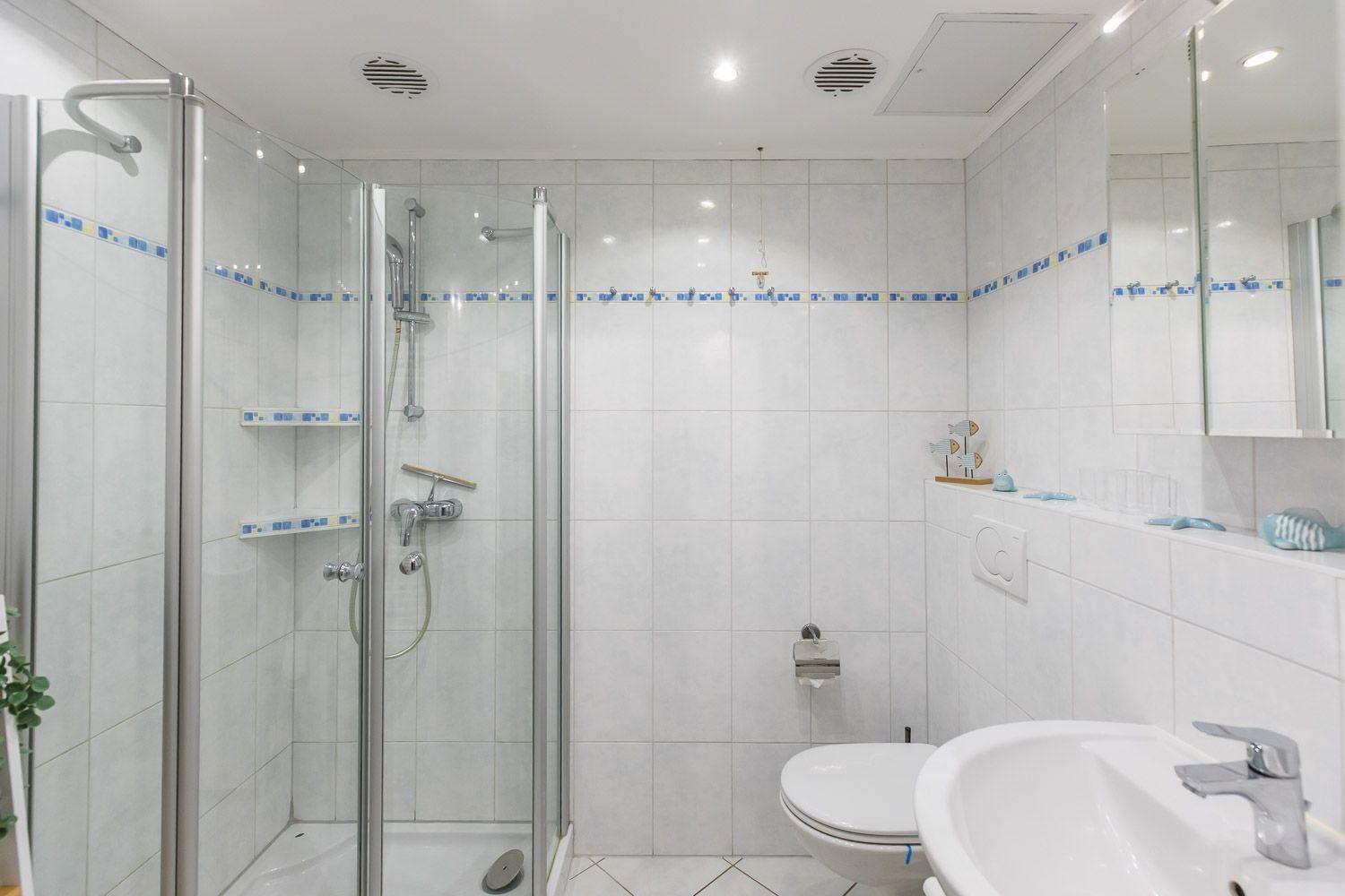 Badezimmer mit Dusche, Toilette und Waschbecken. Weiße Fliesen mit blauer Bordüre.