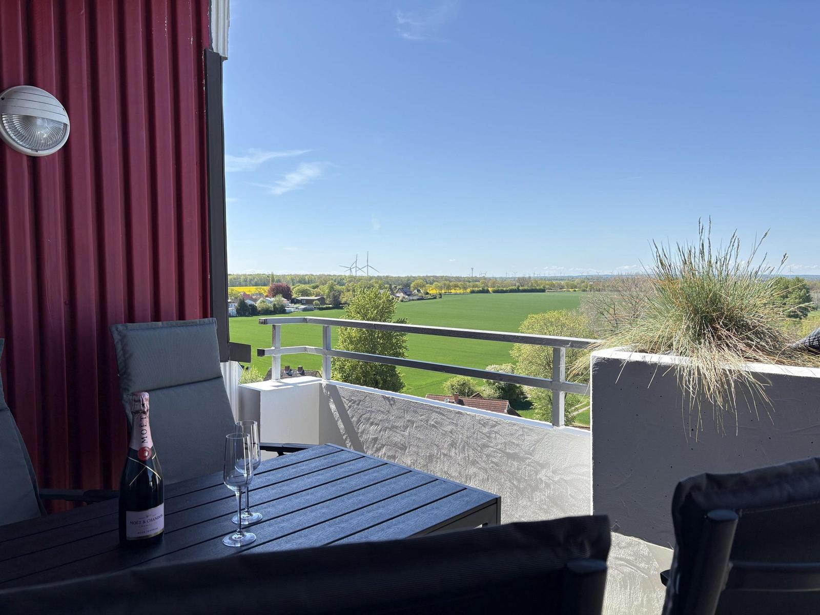 Terrasse mit Tisch, Stühlen und Flasche. Blick auf grüne Felder und Windräder.