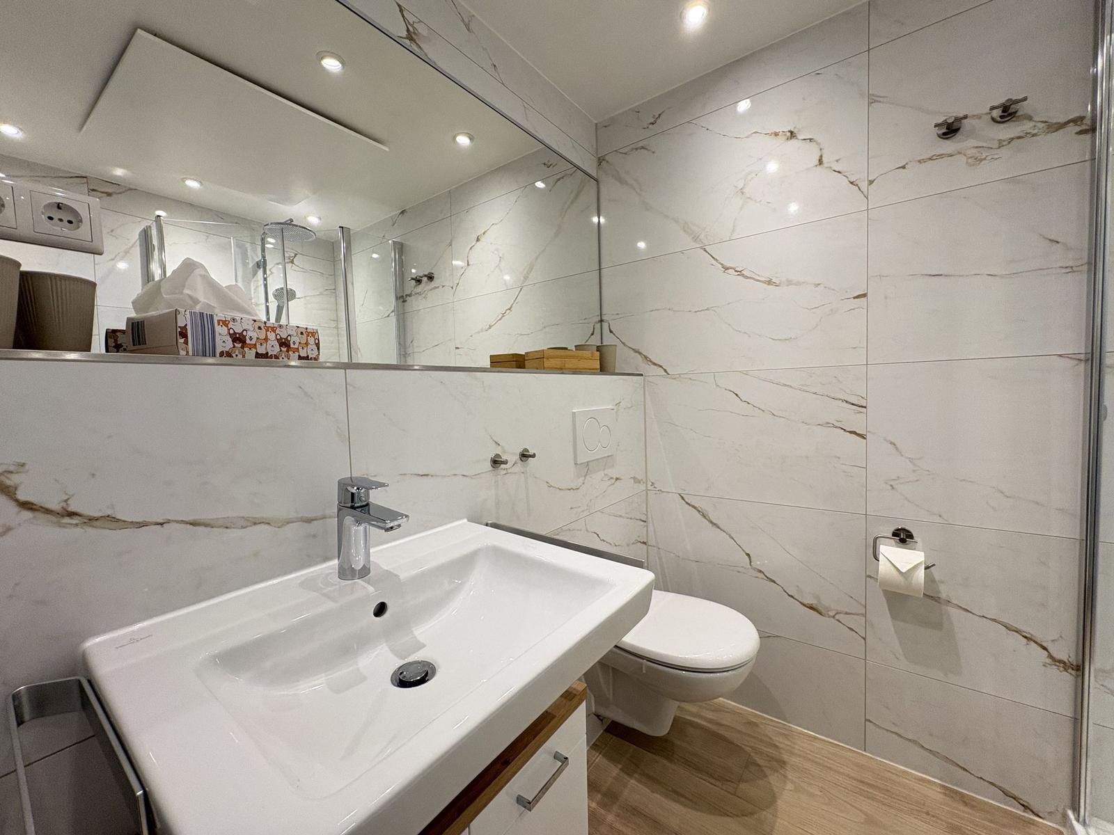 Badezimmer mit Waschbecken, Toilette und Dusche. Wandfliesen im Marmoroptik.
