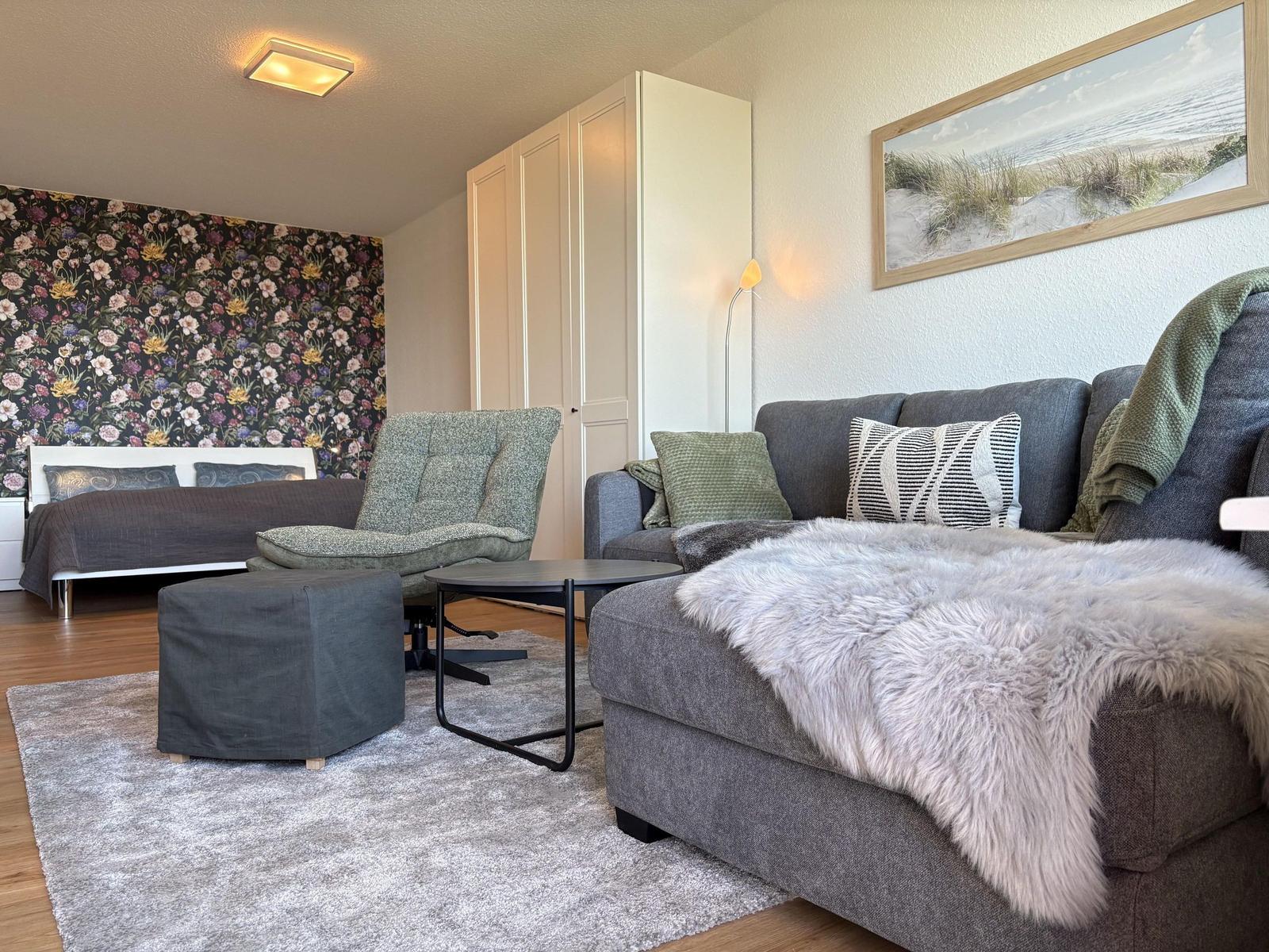 Wohnzimmer mit Sofa, Sessel und Bett. Wand mit Blumenmotiv. Teppich und Decke.