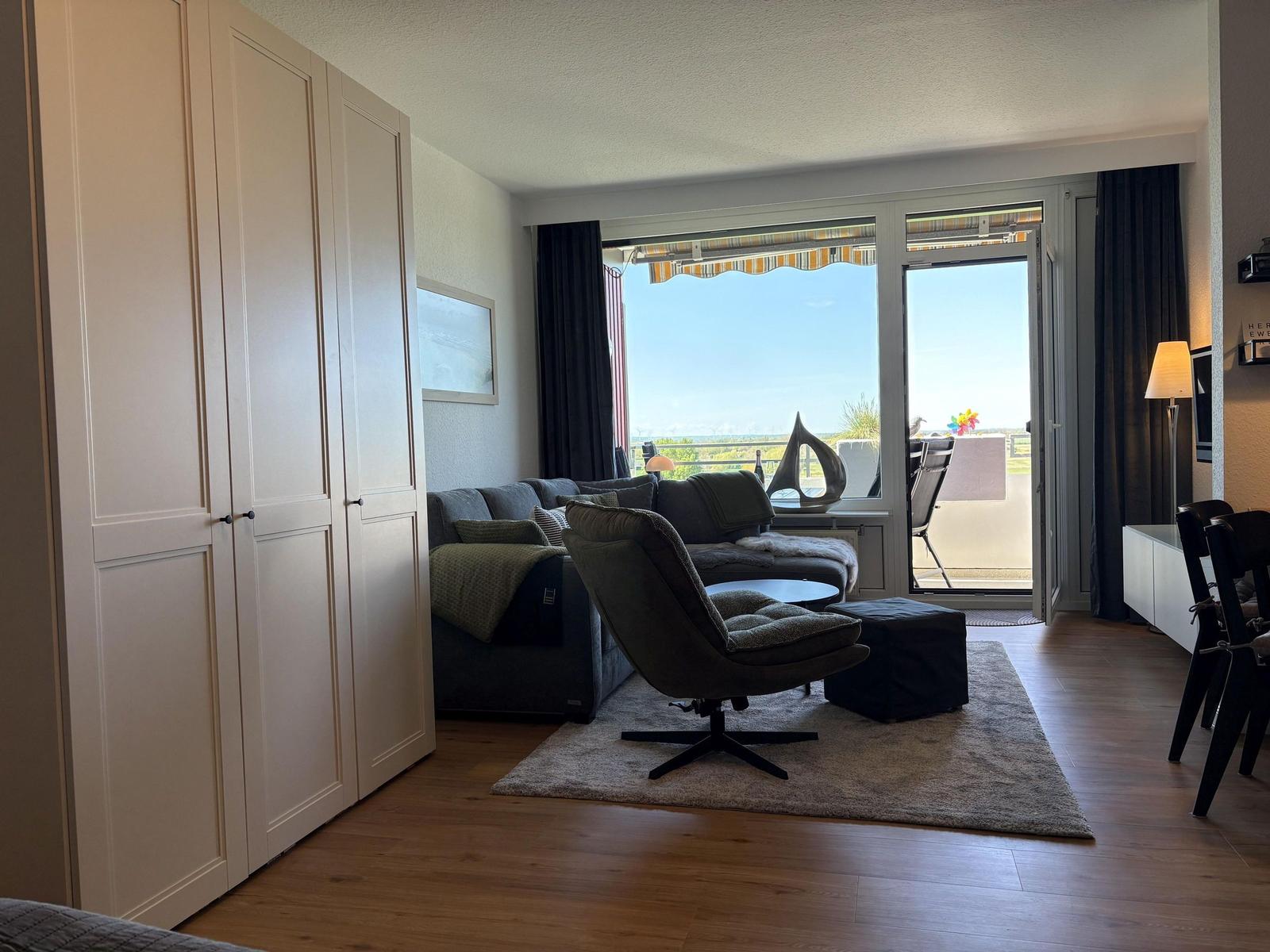 Wohnzimmer mit Sofa, Sessel und Balkon. Holzboden und weiße Schrankwand.