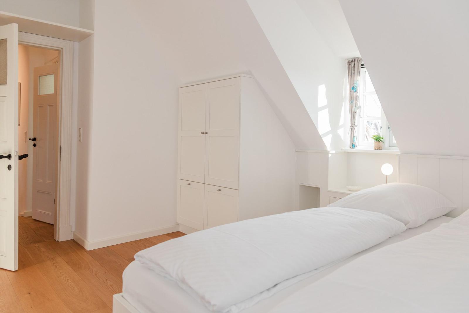 Schlafzimmer mit weißem Bett, Kleiderschrank und Dachschrägenfenster.