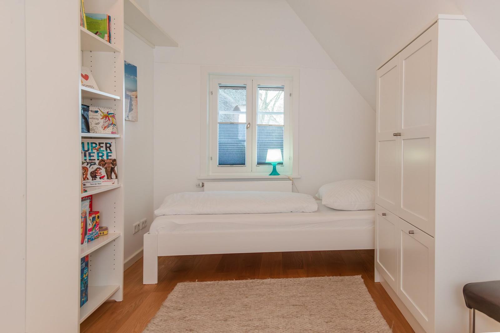 Schlafzimmer mit weißem Bett, Bücherregal und Kleiderschrank unter schrägem Dach