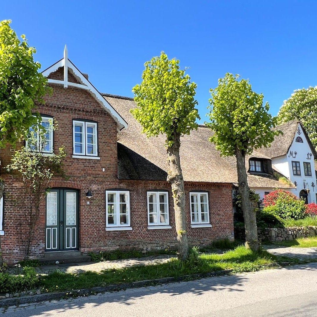 Rotes Backsteingebäude mit strohgedecktem Dach und weißen Fenstern. Grüne Bäume und ein gepflasterter Weg.