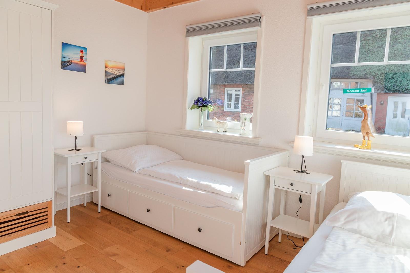 Zimmer mit zwei Betten, weißen Nachttischen und Fenstern mit Blick auf Straße.