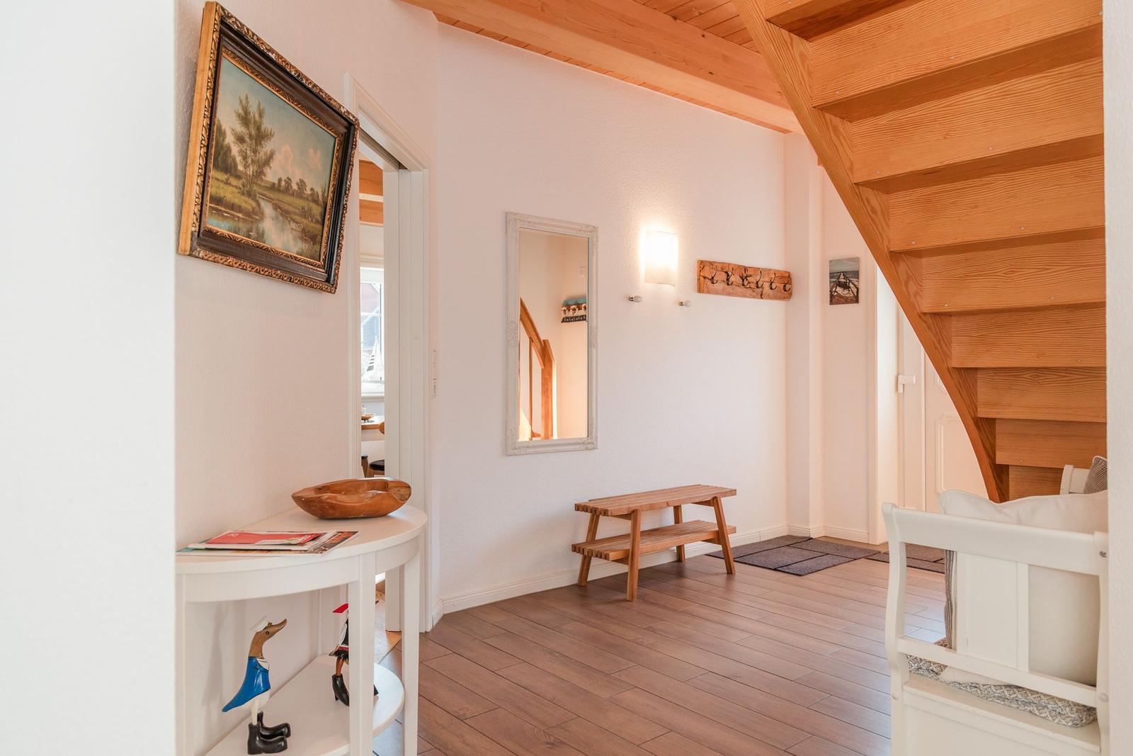 Eingangsbereich mit Holztreppe, Bänkchen und Wandspiegel. Holzdielenboden und weiße Wände.