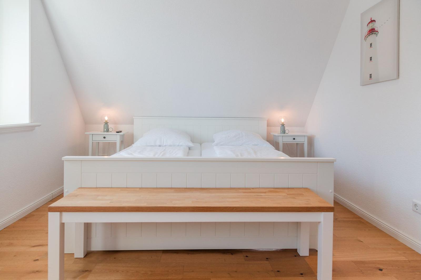 Schlafzimmer mit weißem Bett, Holzbank und Nachttischen unter schrägen Wänden.