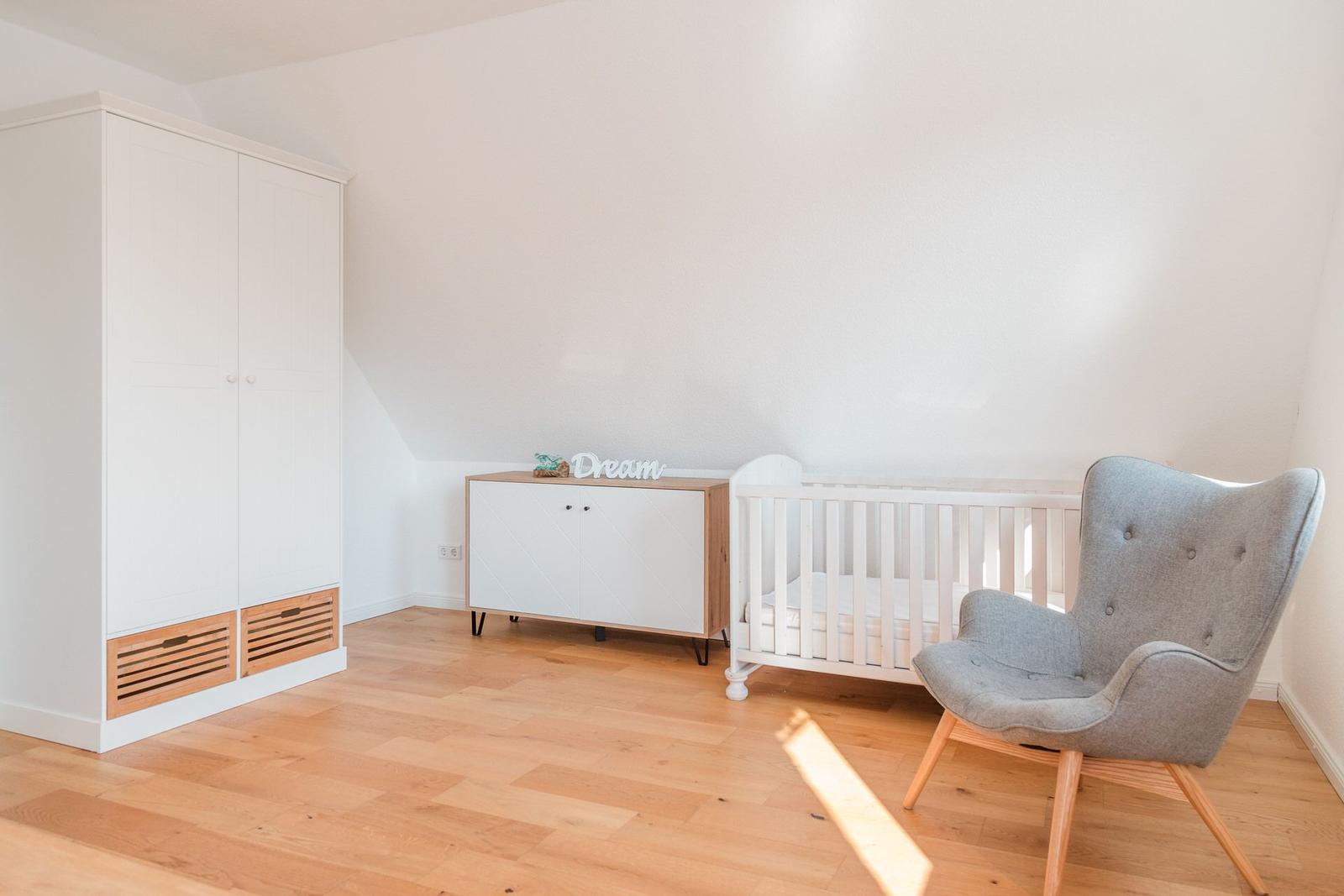 Kinderzimmer mit weißem Schrank, Wickelkommode und Kinderbett. Holzboden und graue Sessel.