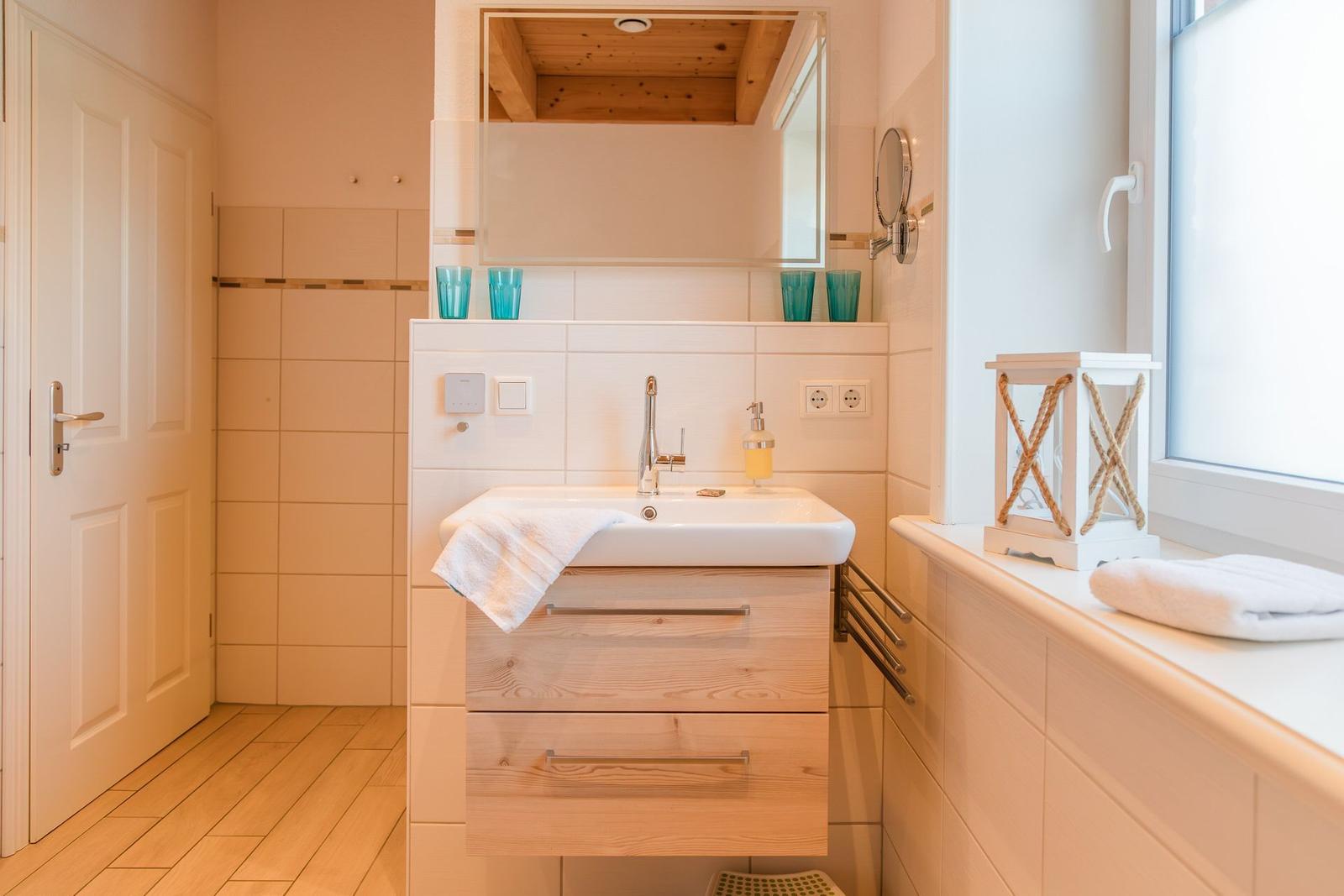 Badezimmer mit Waschbecken, Spiegel und Fenster. Holzschrank und Handtuch auf Fensterbank.