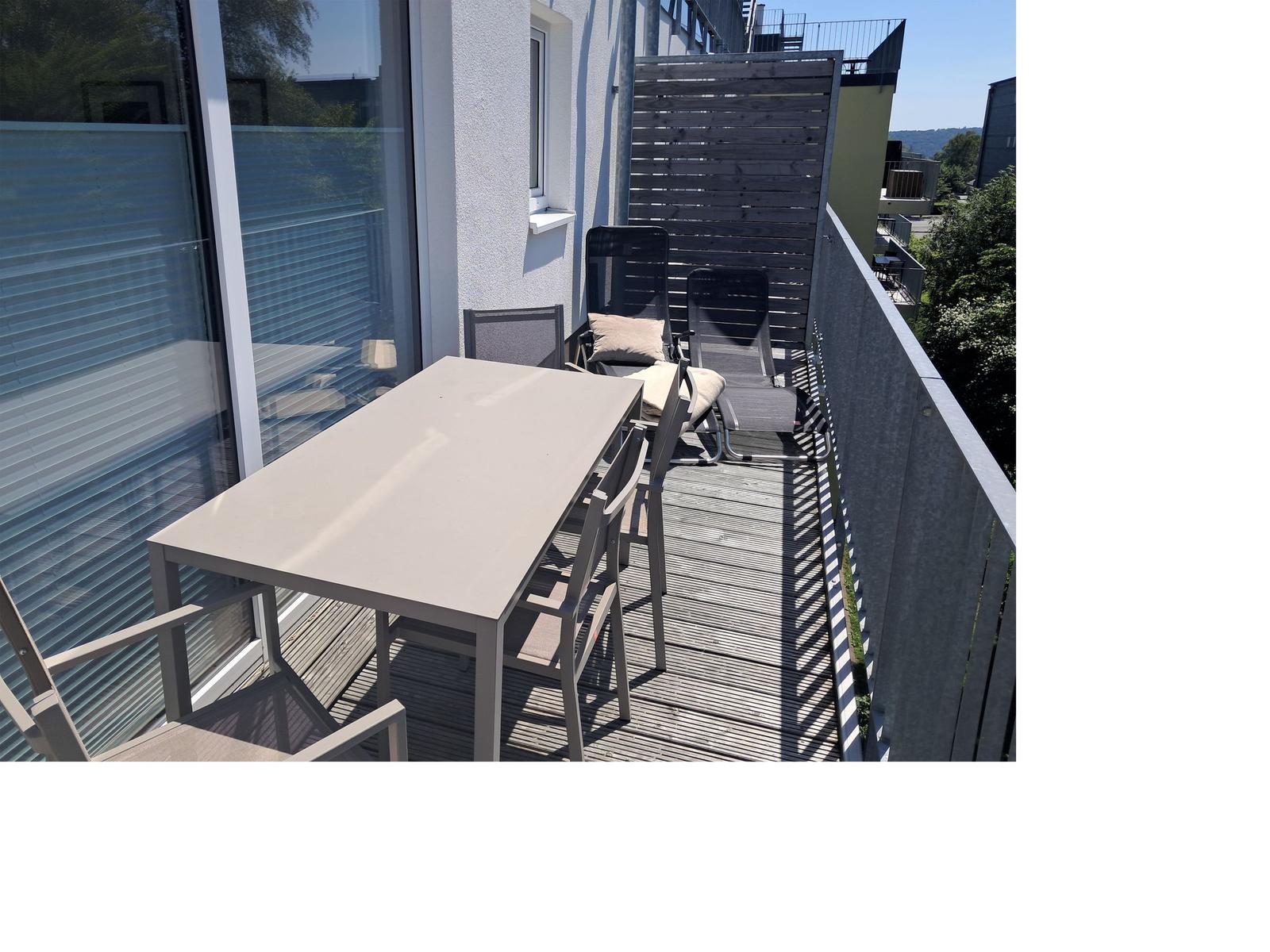 Terrasse mit Tisch, Stühlen und Liegen. Holzdecke und Sicht auf Grünflächen.