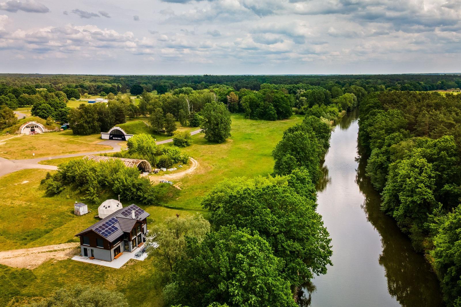 Haus mit Solaranlage neben Fluss und Wald. Hangar-ähnliche Strukturen im Hintergrund.
