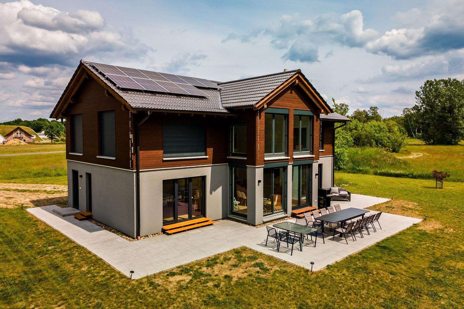 Modernes Haus mit Solaranlage, Terrasse und Garten. Große Fenster und Holzverkleidung.