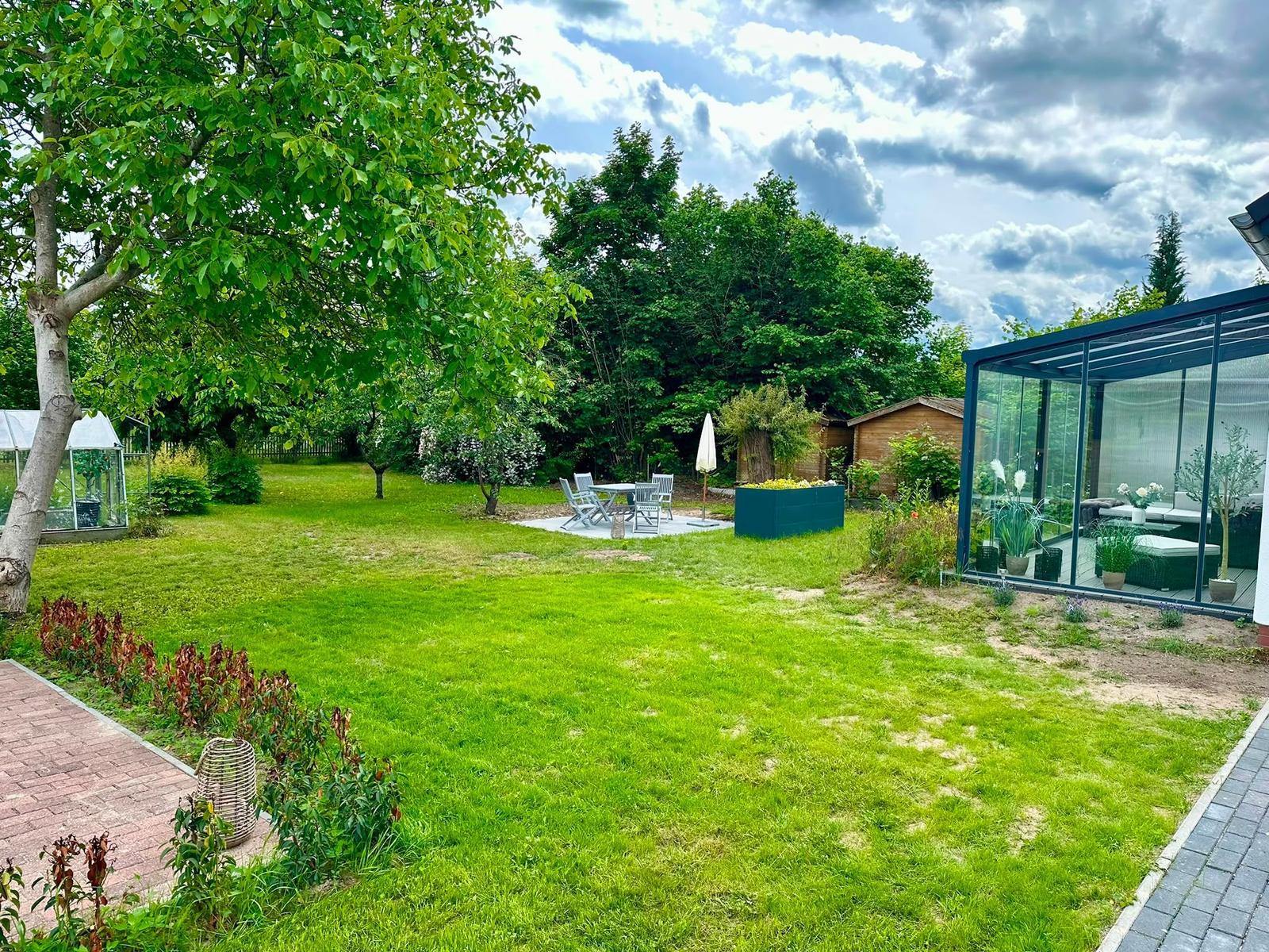 Großer Garten mit Terrasse, Sitzgruppe und Glasbau. Grünfläche und Bäume umgeben den Außenbereich.