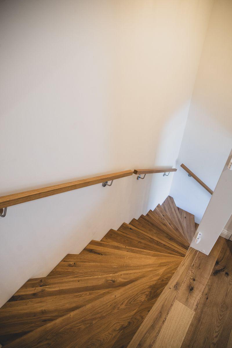 Holztreppe mit Handlauf führt nach oben. Wandfarbe weiß. Bodenbelag aus Holz.