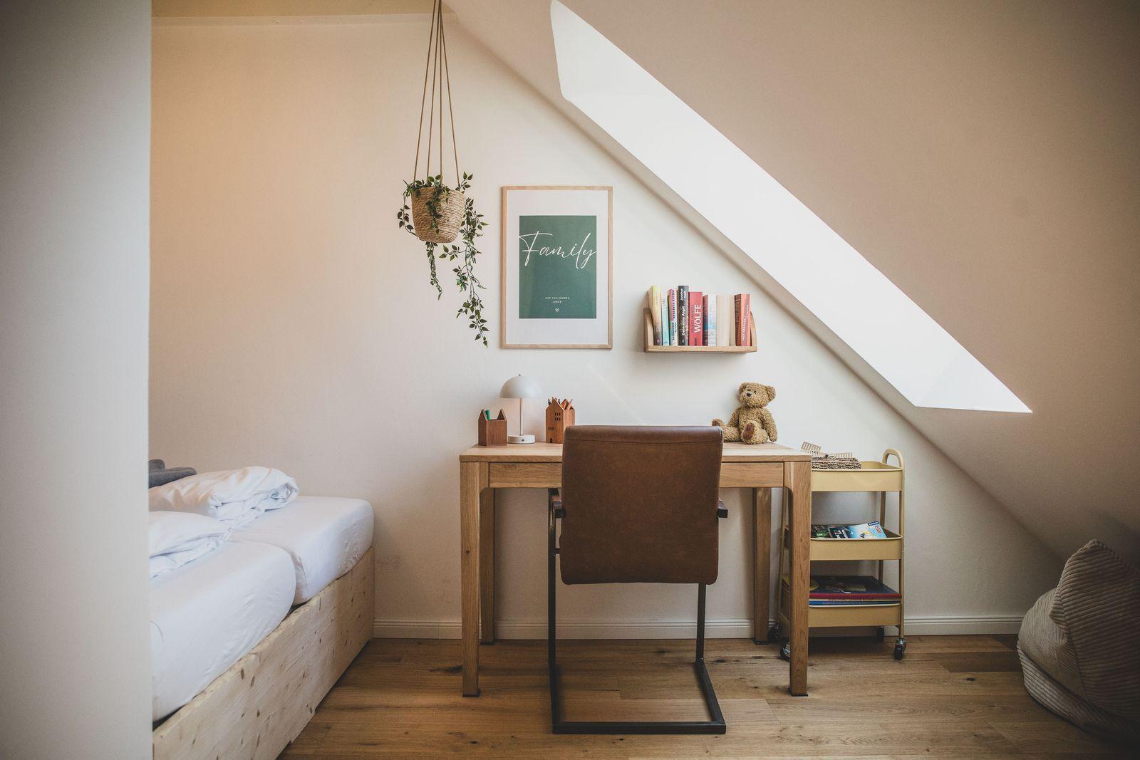 Ein Schlafzimmer mit Schreibtisch, Bett und Dachschräge. An der Wand hängt ein Bild und eine Pflanze.