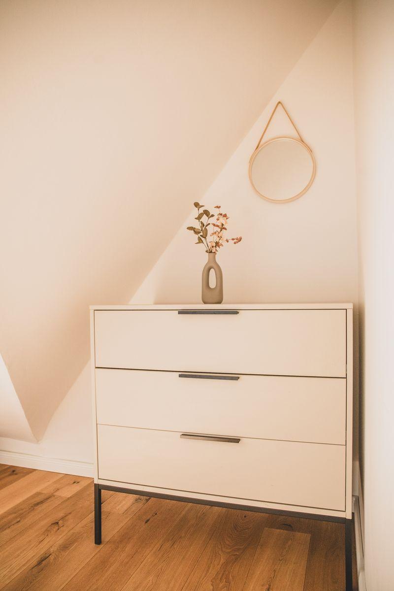Schrank mit drei Schubladen, Vasen mit Blumen und runder Wandspiegel.