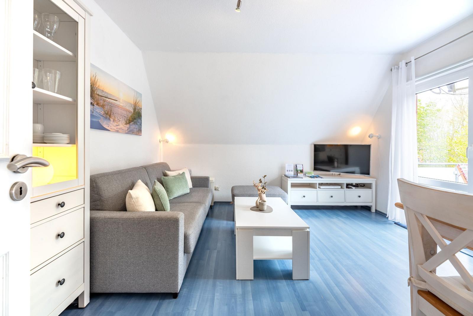 Wohnzimmer mit Sofa, Couchtisch und Fernseher. Blauer Boden und große Fenster.