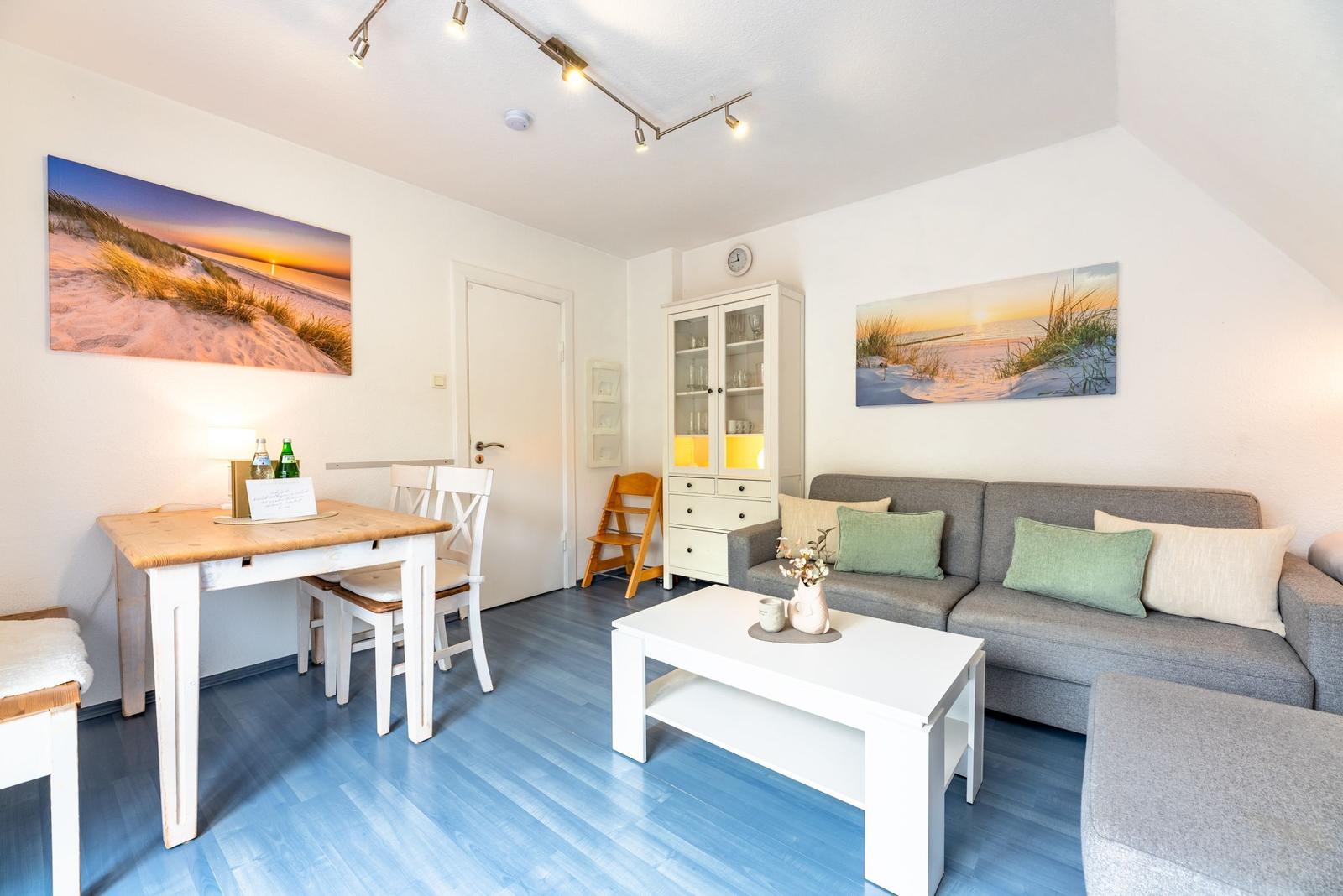 Wohnzimmer mit Sofa, Esstisch und Küchenzeile. Blauer Boden, Wandbilder und weißer Schrank.