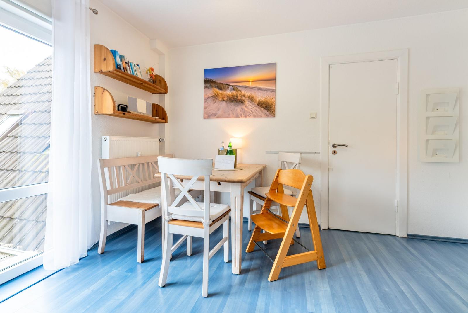 Essbereich mit Tisch, Stühlen und Wandbild. Blauer Boden und Fenster mit Vorhängen.