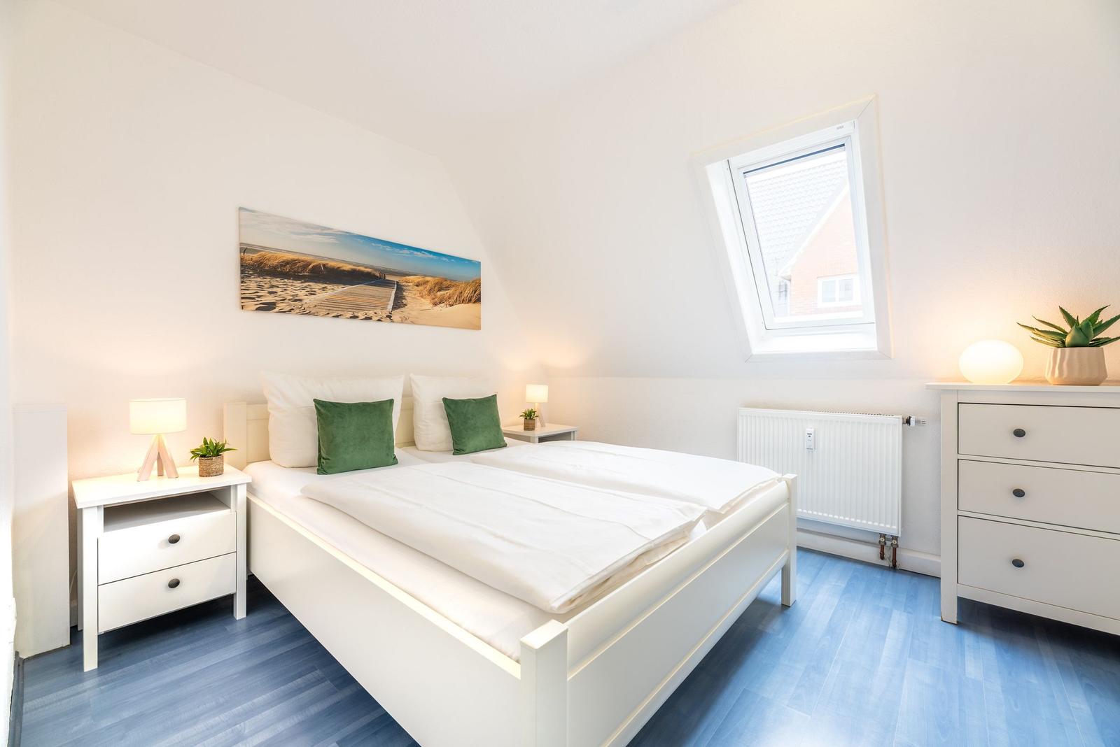 Schlafzimmer mit Doppelbett, Nachttisch und Schrank. Fenster und Wandbild sind sichtbar.