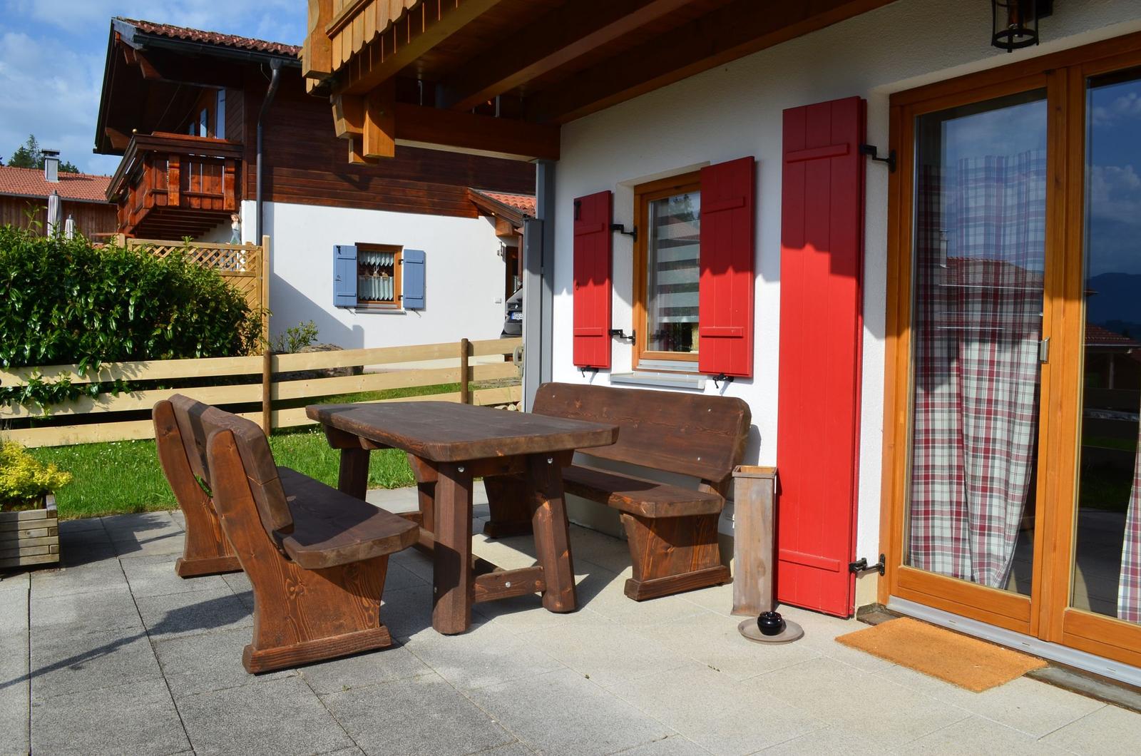 Terrasse mit Holztisch und Bänken, rote Fensterläden, Glastür mit kariertem Vorhang.