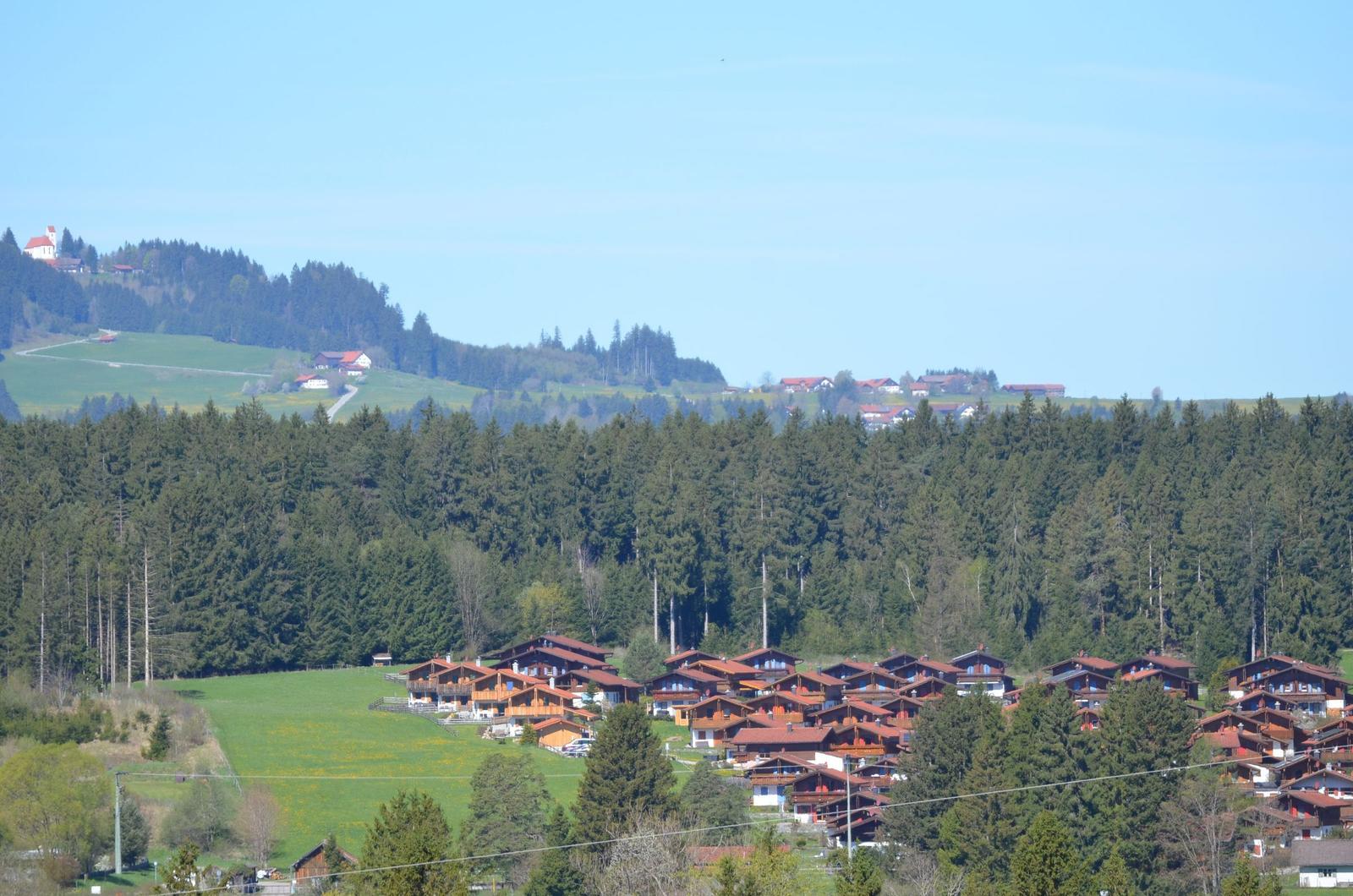 Wohngebiet mit Holzhäusern, umgeben von Wald und Hügeln unter blauem Himmel.