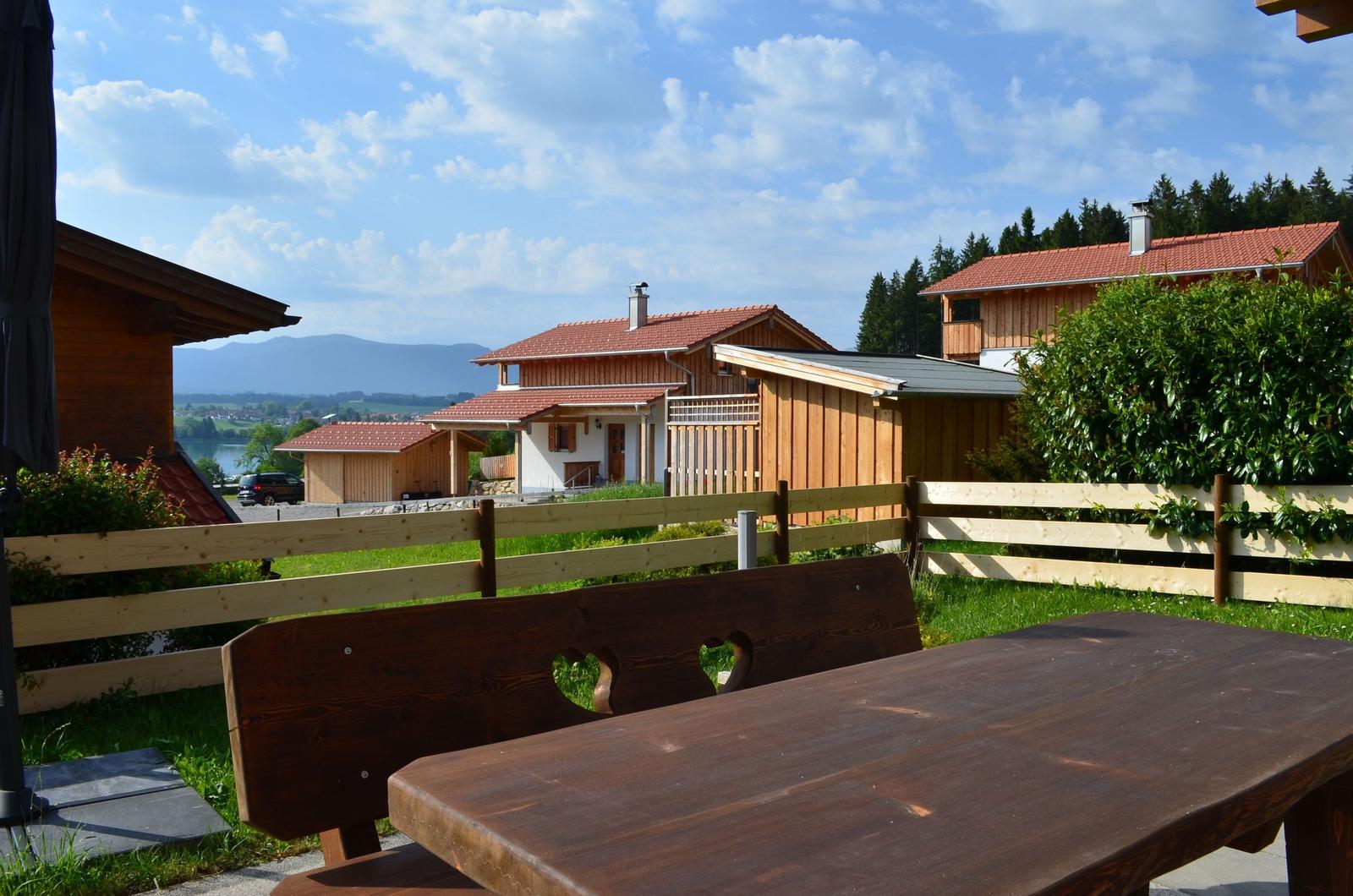 Holztisch und -bank mit Herzöffnungen im Garten. Holzhäuser, Zaun und Berge im Hintergrund.