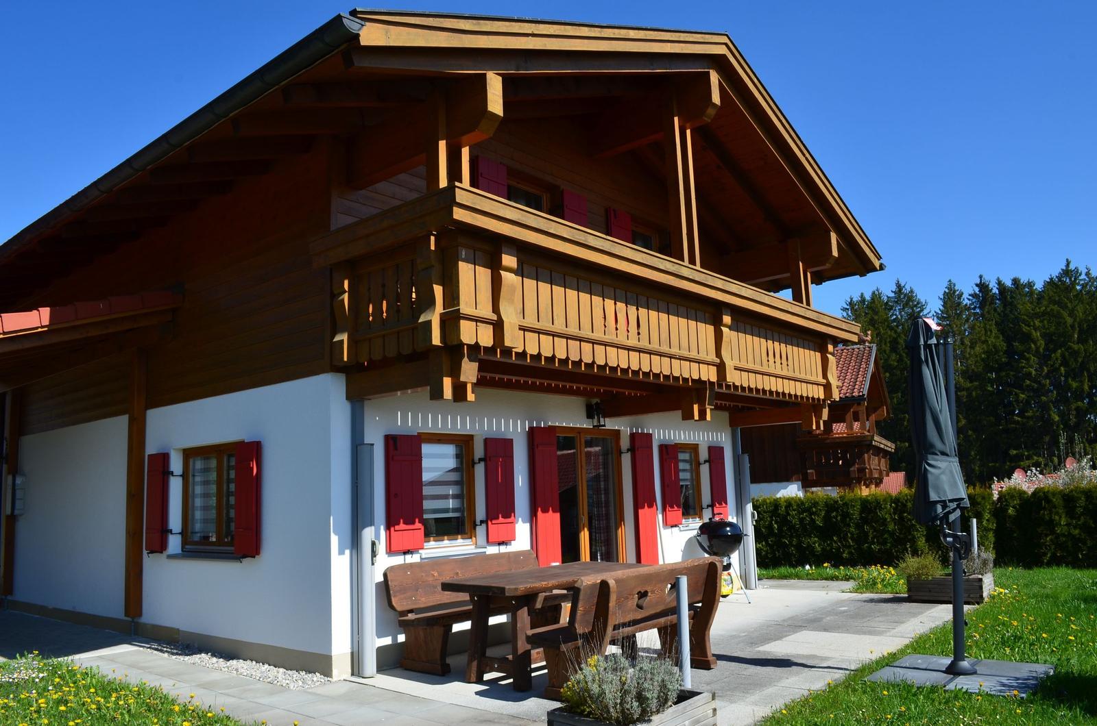 Holzhaus mit Balkon, roten Fensterläden und Gartenmöbeln. Grill und Sonnenschirm vorhanden.