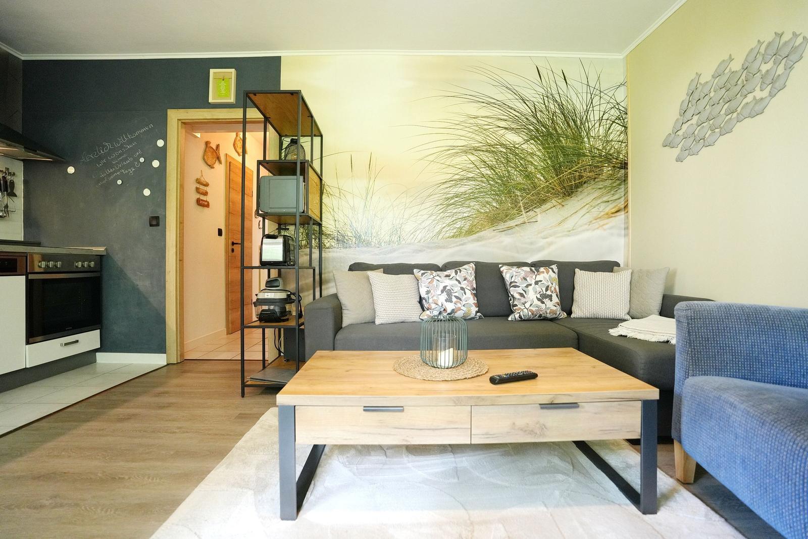 Wohnzimmer mit Sofa, Couchtisch und Küchenbereich. Wandbild mit Strandmotiv.