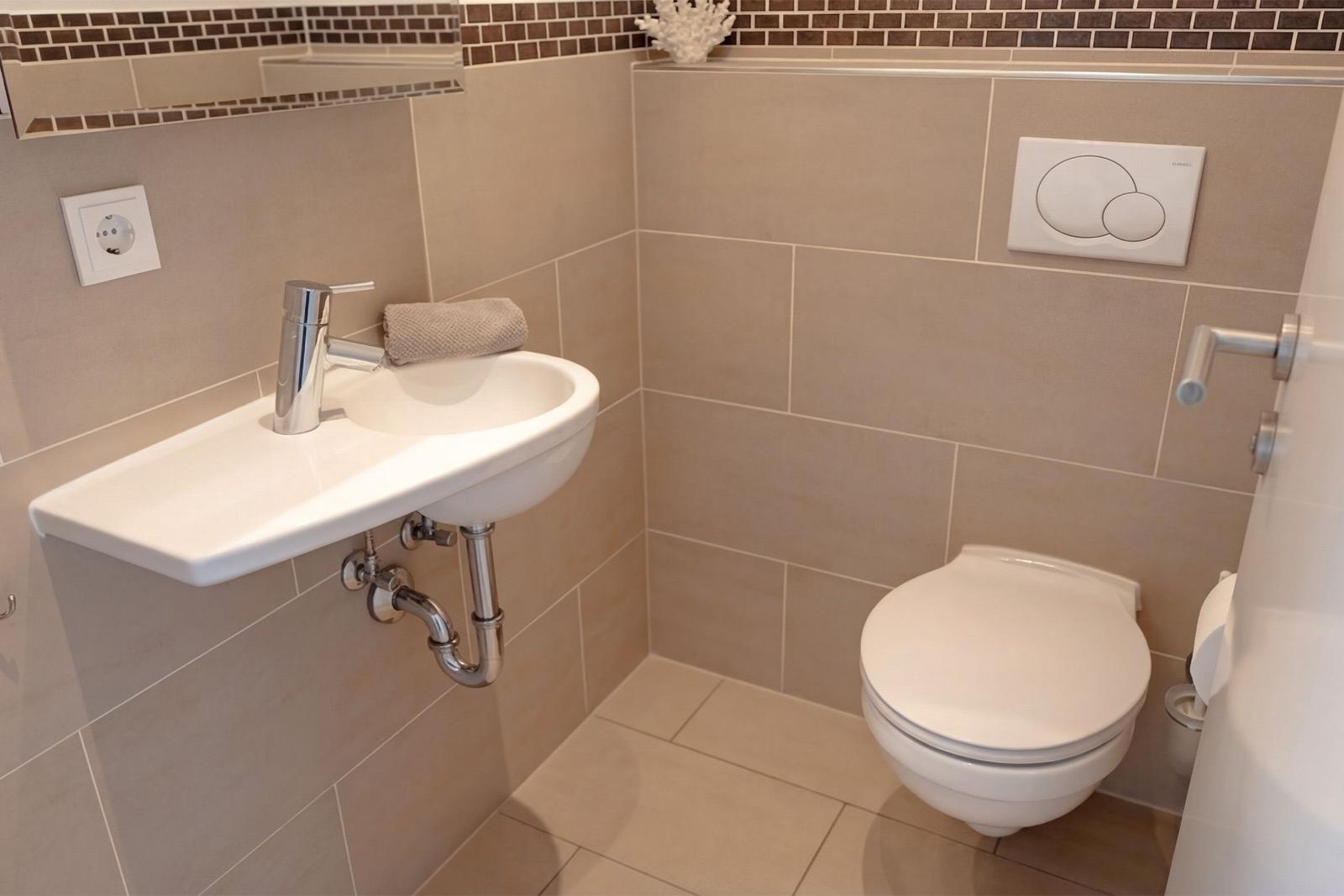 Badezimmer mit Waschbecken, Spiegel und WC. Wandfliesen und Metallarmatur sichtbar.