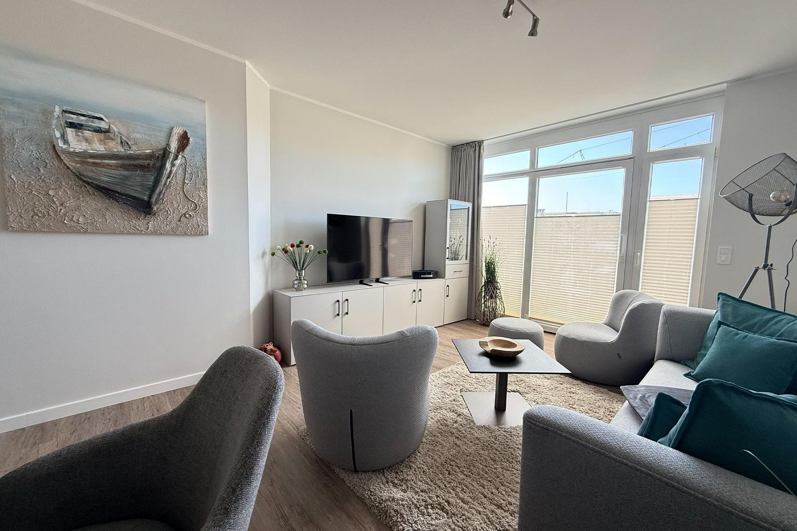 Wohnzimmer mit Sofa, Sesseln und Fernseher. Große Fenster und Wandbild mit Boot.