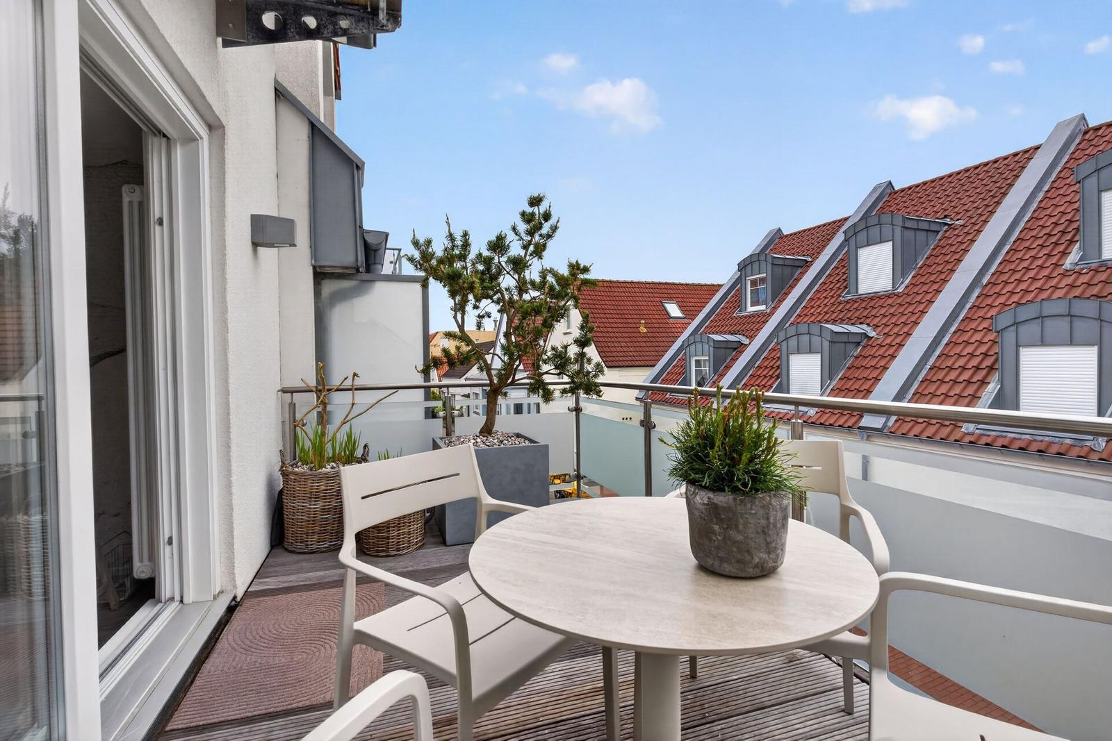 Terrasse mit Tisch, Stühlen und Pflanzen. Blick auf rote Dächer und blauen Himmel.