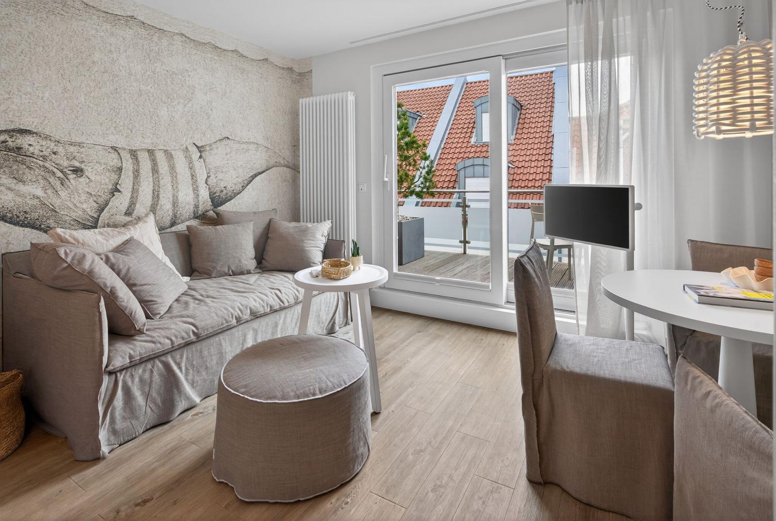 Wohnbereich mit Sofa, Tisch und Stühlen. Wandbild zeigt Wal. Balkon mit Aussicht auf Dächer.