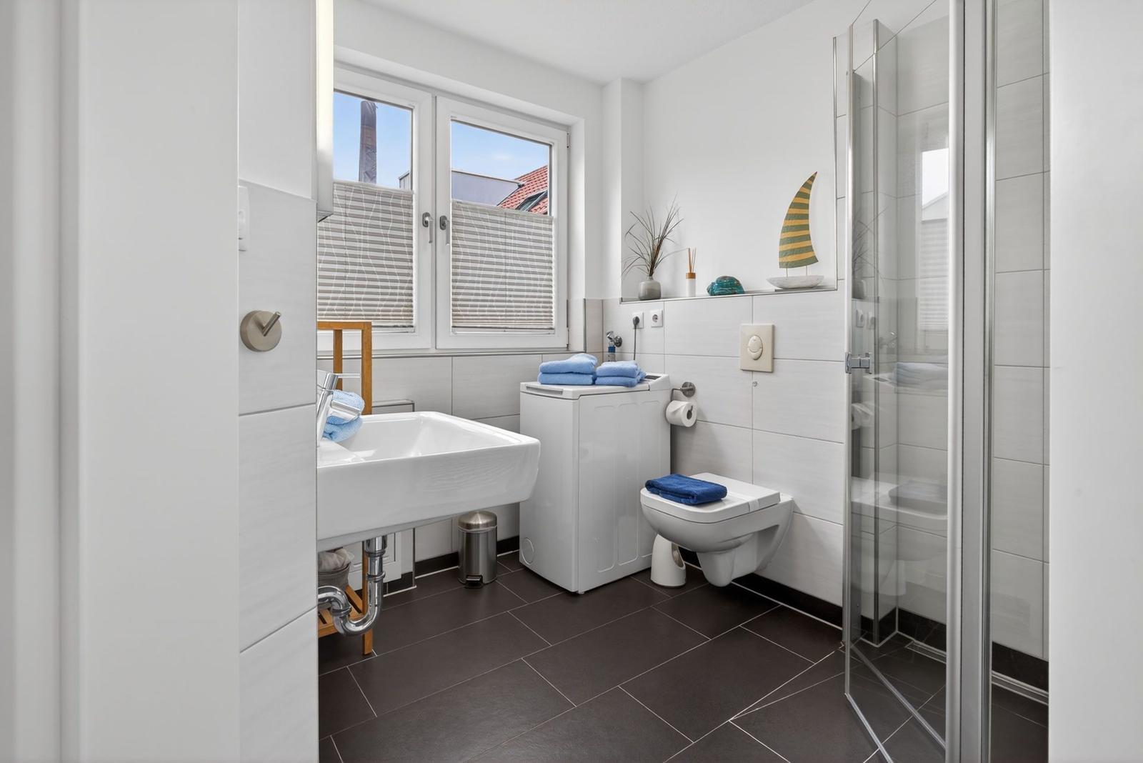 Badezimmer mit Dusche, Toilette, Waschmaschine und Fenster.