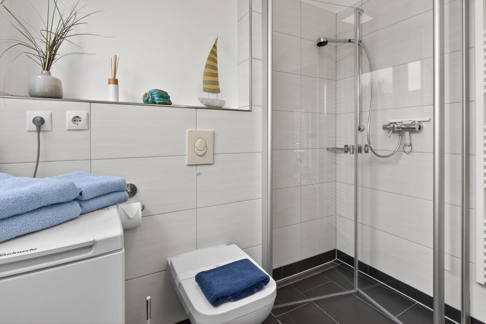 Badezimmer mit Dusche, Toilette und Waschmaschine. Auf der Ablage stehen dekorative Gegenstände.