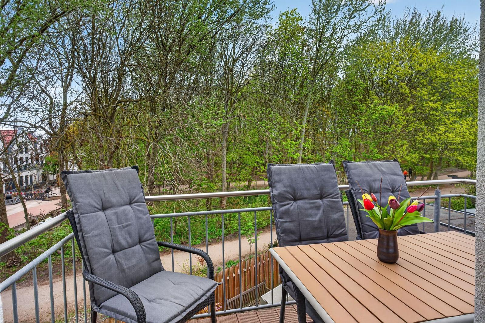 Terrasse mit Tisch, drei Stühlen und Blumen. Blick in einen grünen Park.