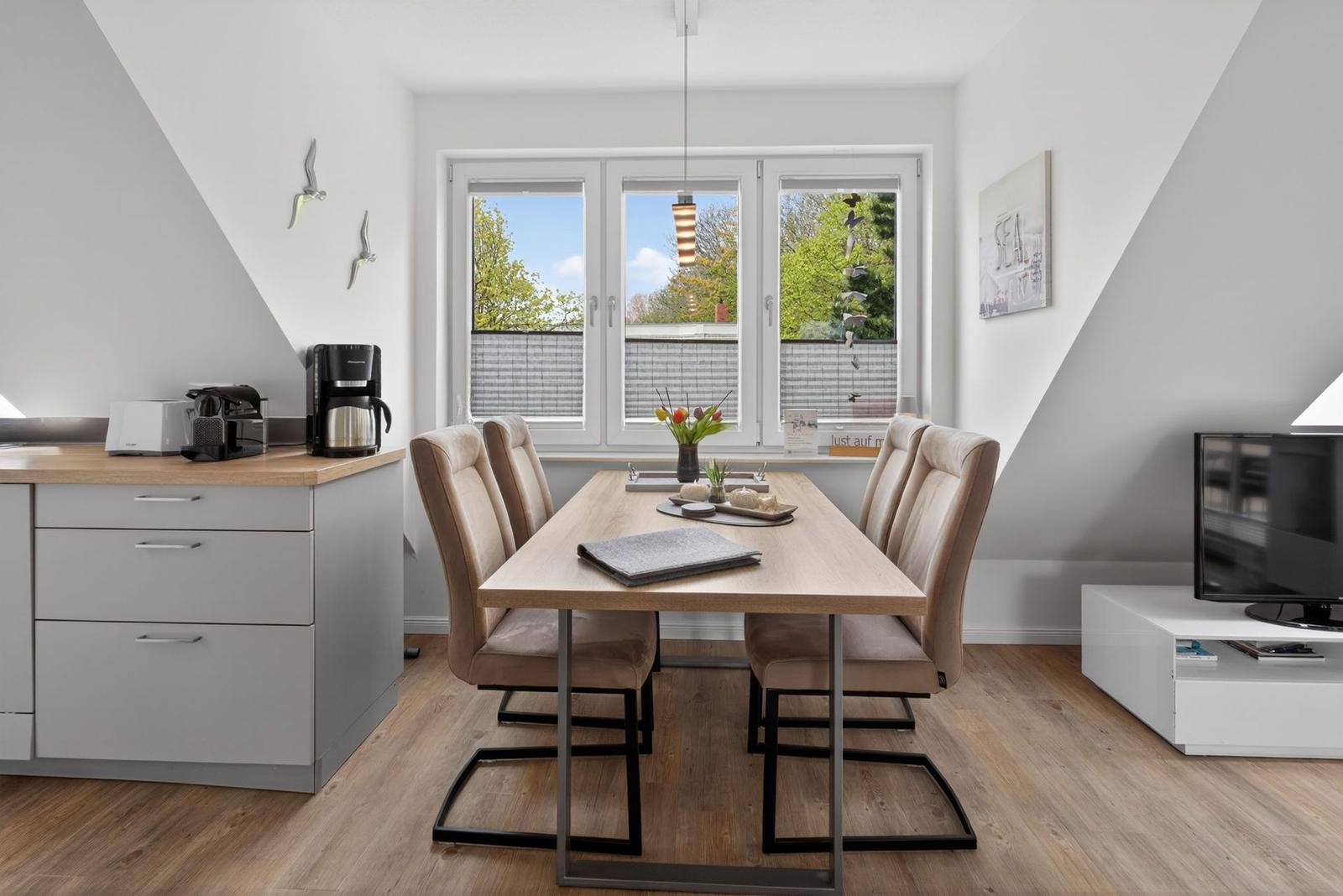 Esszimmer mit Tisch, Stühlen und Fenster. Küchenzeile und Fernseher im Hintergrund.