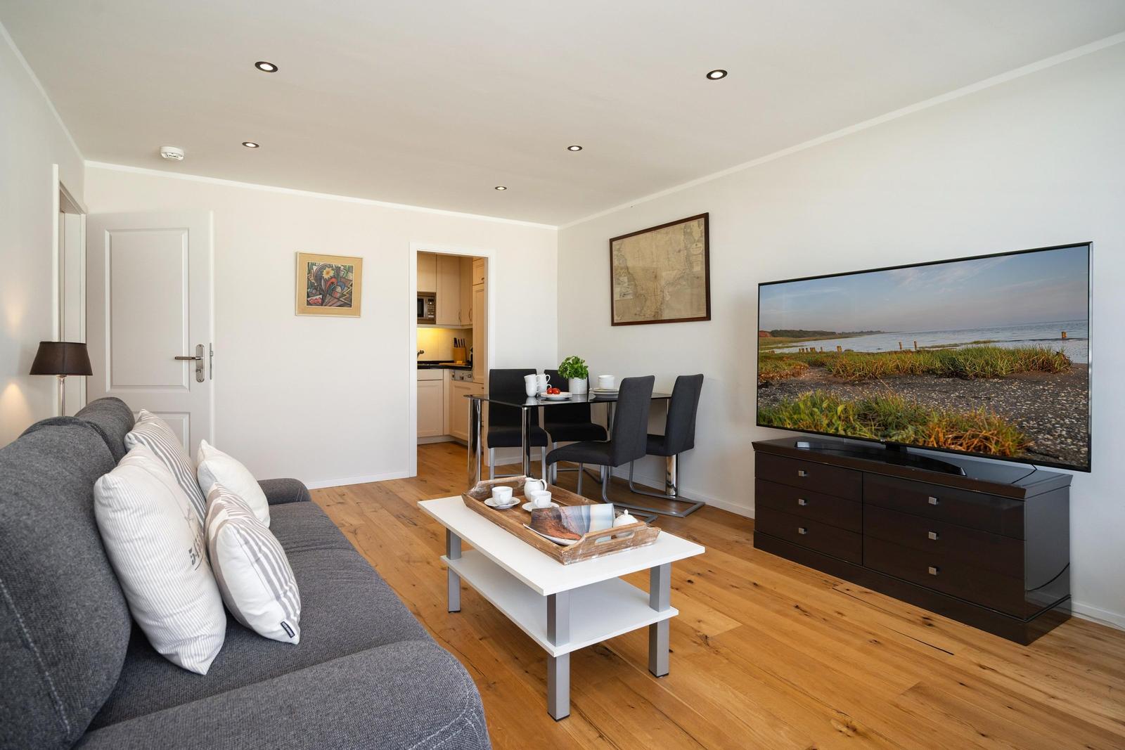 Wohnzimmer mit Sofa, Couchtisch, TV und Essbereich. Holzboden und weiße Wände.