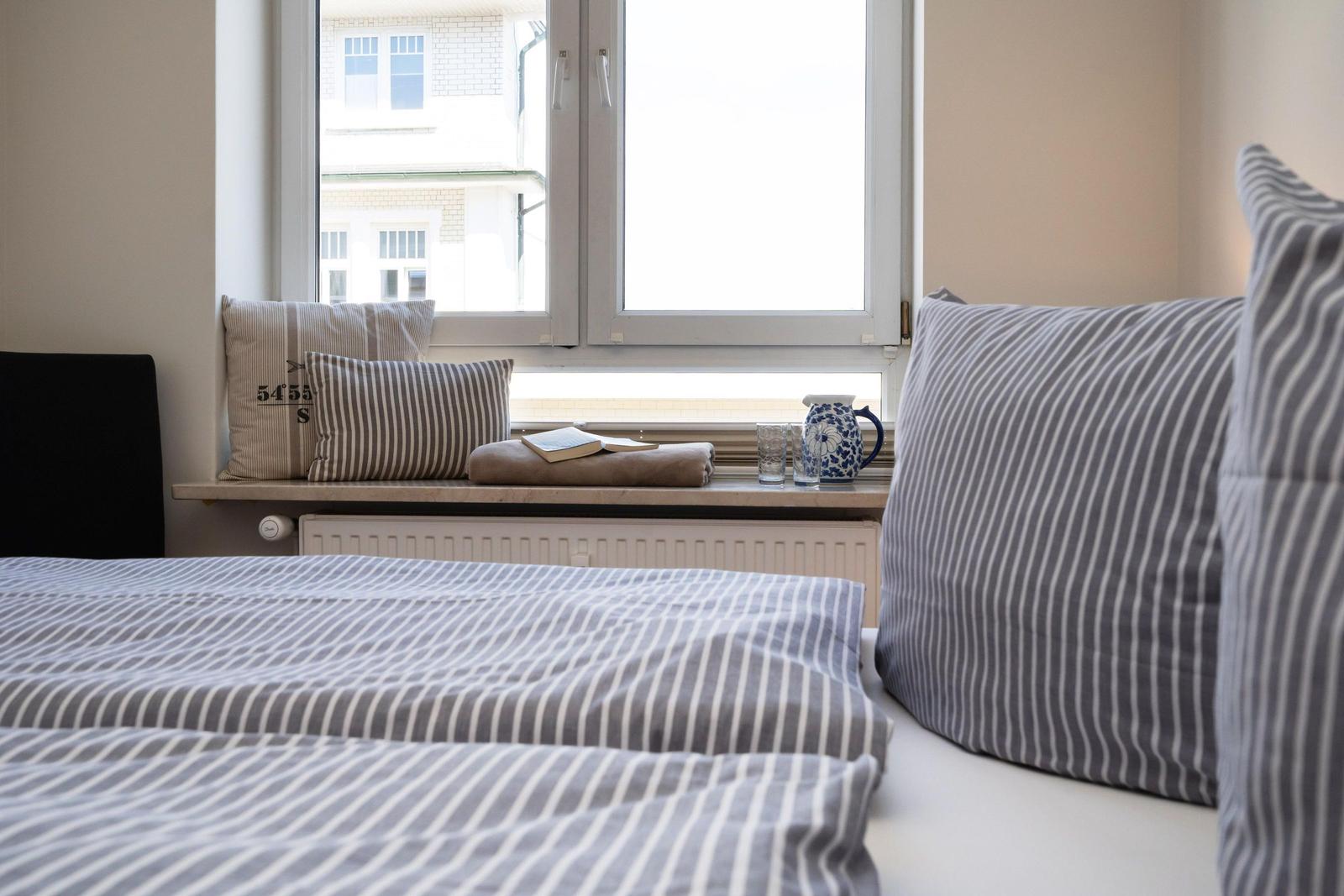 Ein Bett mit gestreiften Kissen und einer Decke. Auf dem Fensterbrett steht eine Teekanne und ein Buch.