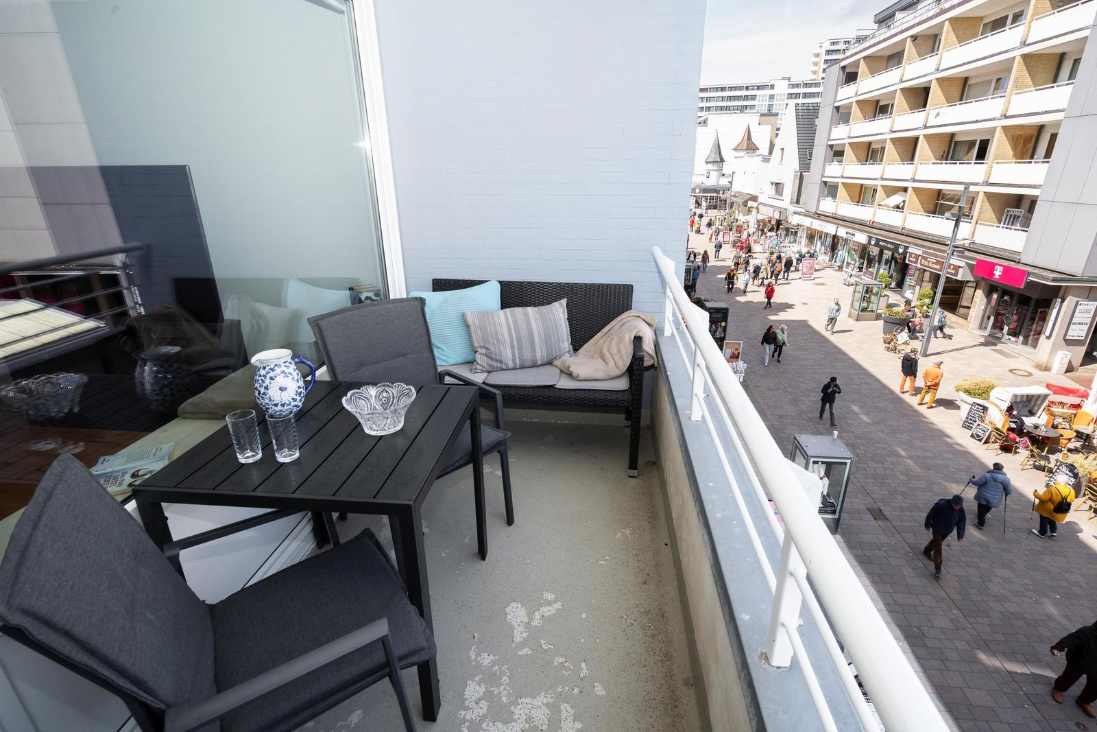 Terrasse mit Sitzgruppe und Tisch. Blick auf belebte Straße mit Geschäften und Passanten.
