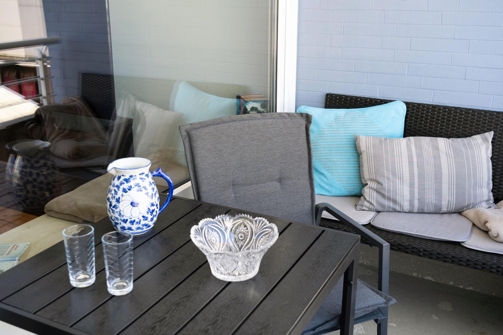Terrasse mit Tisch, Stühlen und Kissen. Auf dem Tisch stehen ein Wasserglas und eine Schale.