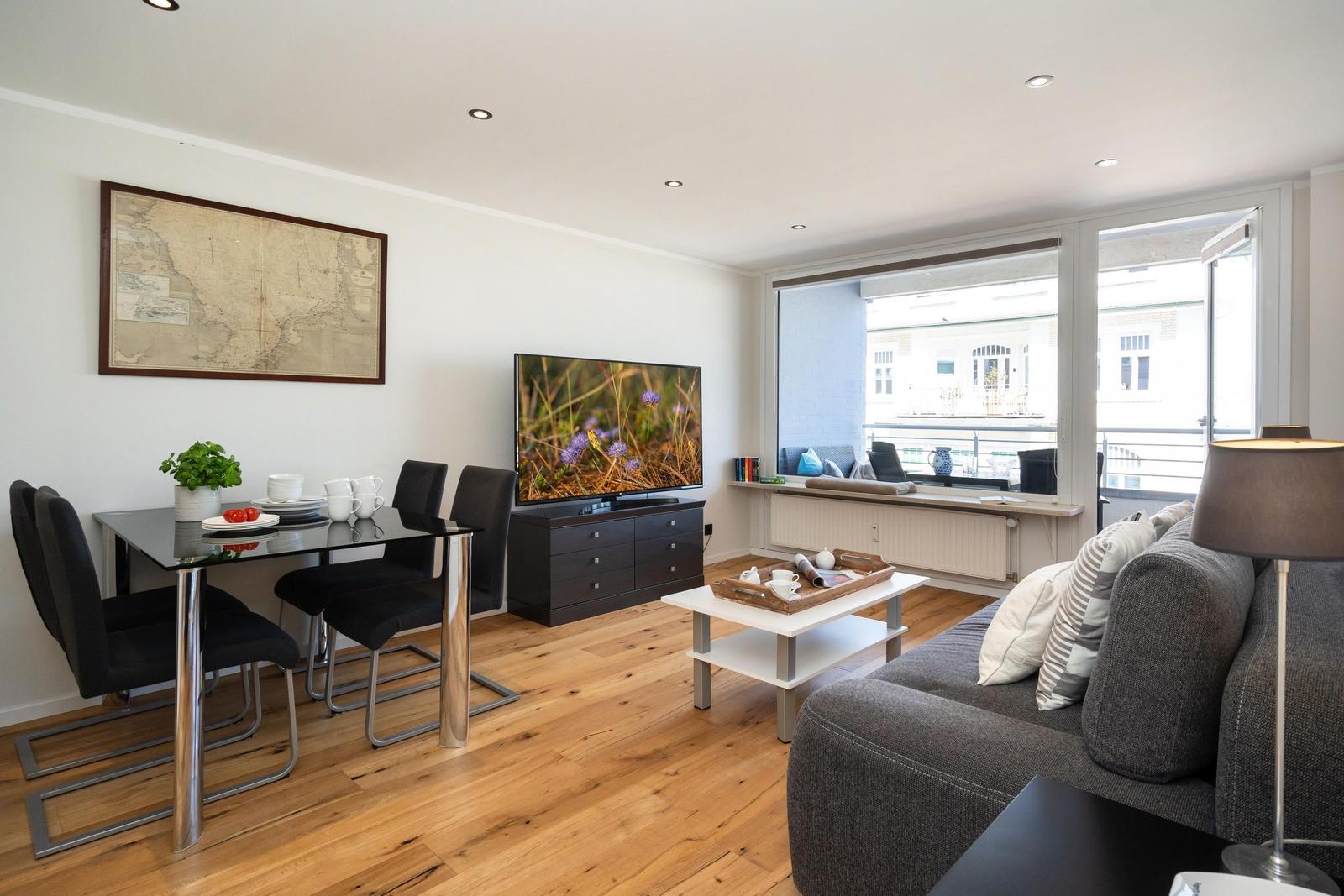 Wohn- und Essbereich mit TV, Sofa und Tisch. Holzboden und große Fenster.